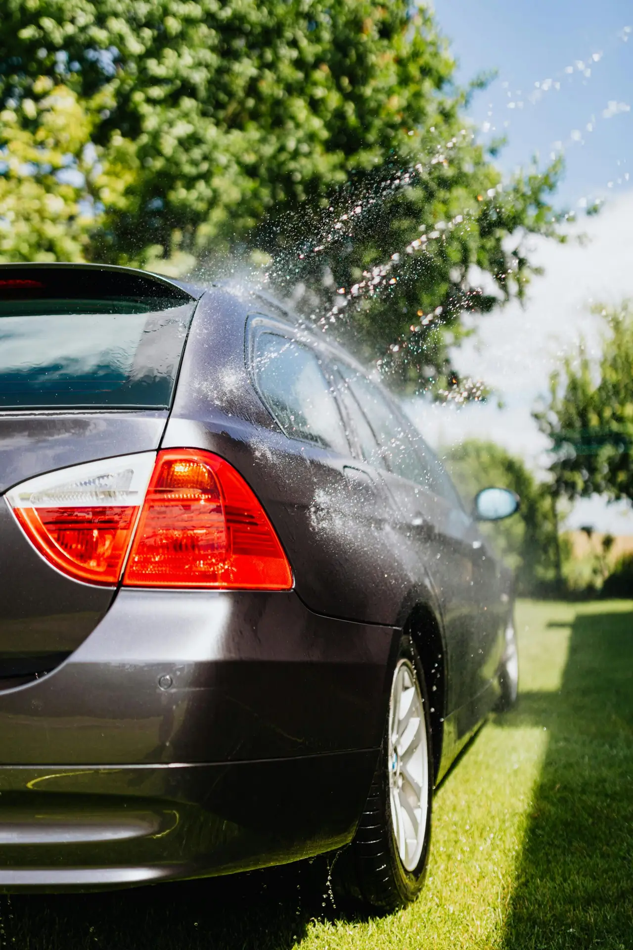 Rear view of a silver car being cleaned with water splashes on a sunny day.