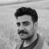 A monochrome portrait of a young man with a mustache, outdoors in a field.